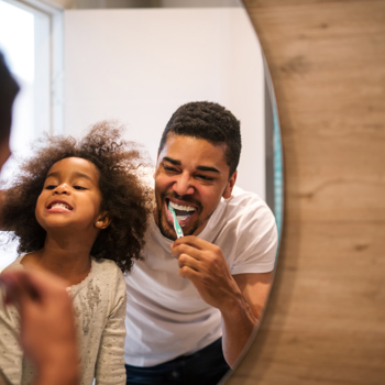 A father laughing in the mirror while brushing his teeth with his young child.