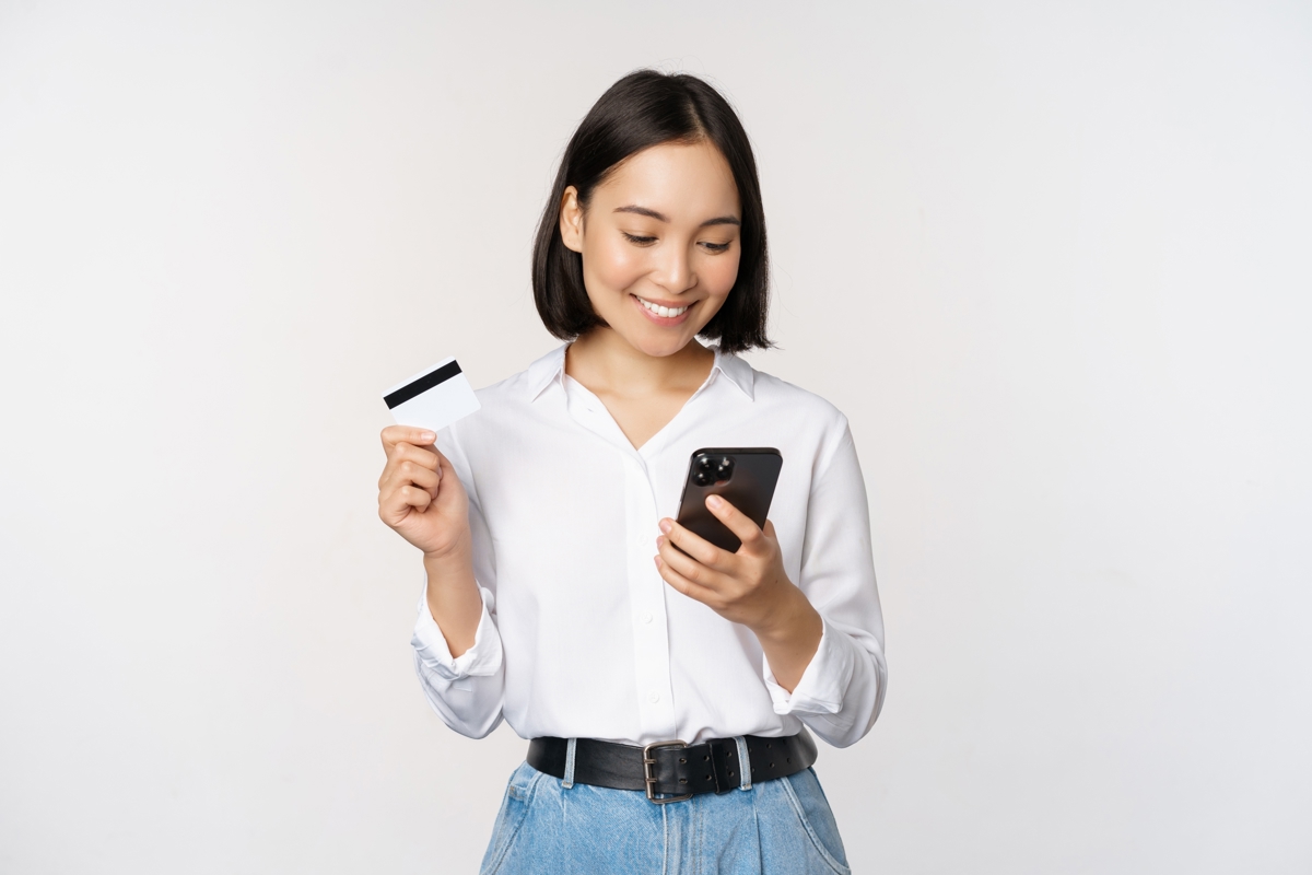 A smiling woman holding a credit card and looking at her phone with a white background.